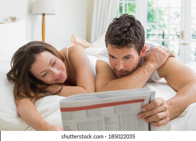 Relaxed Young Couple Reading Newspaper In Bed At Home