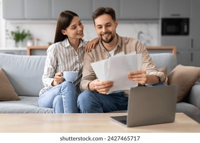 Relaxed young couple reviewing documents with smiles, woman holding cup of coffee, both sitting by laptop on cozy couch at home in living room - Powered by Shutterstock