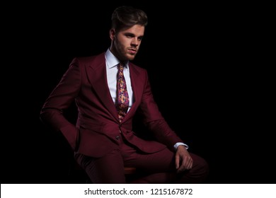 Relaxed Young Elegant Man In Burgundy Suit Is Resting On A Chair On Black Background