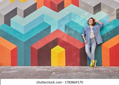 Relaxed young female in denim outfit smiling and looking away while leaning on multicolored graffiti wall with geometric ornament on city street