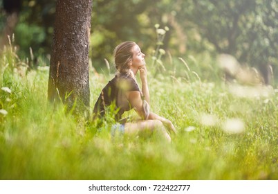 Relaxed young girl sitting under tree on meadow at summer day