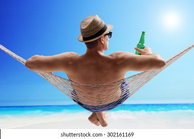 Relaxed young guy lying in a hammock and drinking beer on a sunny beach by the ocean