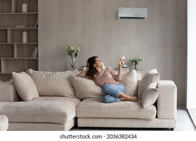 Relaxed young hispanic female homeowner sitting on huge comfortable couch, turning on air conditioner with remote controller, switching on cooler system, setting comfortable temperature in living room - Shutterstock ID 2036382434