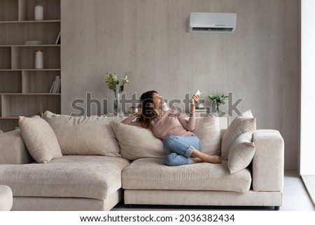 Relaxed young hispanic female homeowner sitting on huge comfortable couch, turning on air conditioner with remote controller, switching on cooler system, setting comfortable temperature in living room