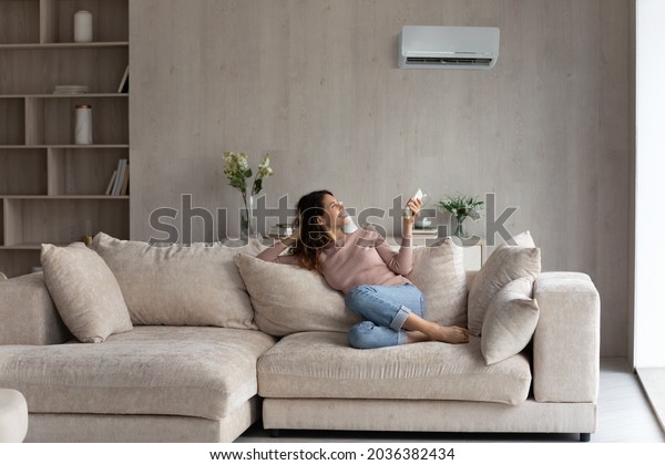 Relaxed young hispanic female homeowner sitting on\
huge comfortable couch, turning on air conditioner with remote\
controller, switching on cooler system, setting comfortable\
temperature in living\
room