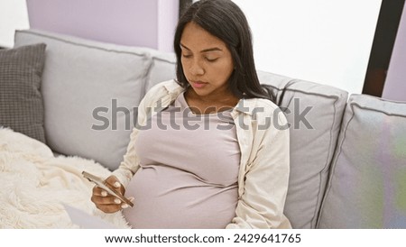 Relaxed young hispanic woman, joyfully wrapped in a blanket, sitting on her home's living room sofa, discloses her pregnancy news online using her smartphone, completely absorbed in motherhood bliss.