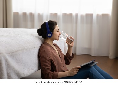 Relaxed young indian ethnicity woman in wireless headphones involved in using digital computer tablet, drinking glass of fresh pure mineral water, enjoying break pause time sitting on floor in bedroom - Powered by Shutterstock