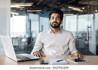 Relaxed young Indian man sitting in office at desk with laptop and documents and resting with closed eyes. - Powered by Shutterstock