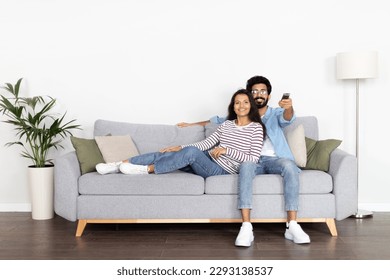 Relaxed young indian spouses sitting on sofa at home, watching movie together, handsome husband hugging his pretty wife, enjoying TV show, holding remote control, copy space, full length