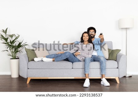 Relaxed young indian spouses sitting on sofa at home, watching movie together, handsome husband hugging his pretty wife, enjoying TV show, holding remote control, copy space, full length