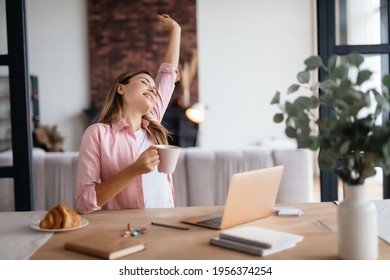 Relaxed Young Lady Holding Coffee Mug And Stretching Her Arm Upwards While Sitting At Working Desk