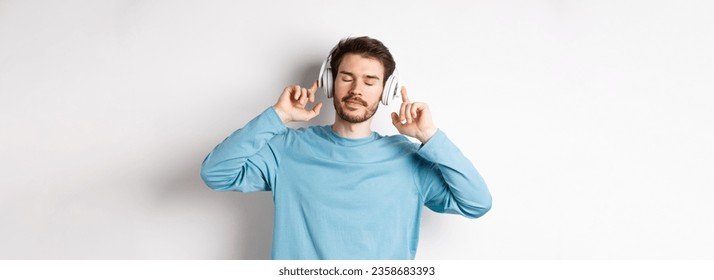 Relaxed young man enjoying favorite song, listening music in headphones with closed eyes and calm face, standing over white background. - Powered by Shutterstock