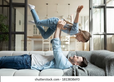 Relaxed Young Man Lying On Cozy Couch, Raising In Air On Straight Arms Laughing Carefree Little Preschool Kid Daughter. Happy Excited Small Child Girl Playing Airplane, Enjoying Weekend With Dad