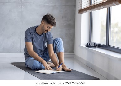 A relaxed young man sits on the floor, writing in a notebook by a large window. He's surrounded by natural light, creating a calm, reflective atmosphere perfect for creativity and focus. - Powered by Shutterstock