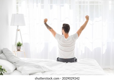 Relaxed young man waking up in bed and stretching his arms, facing the window, back view, free space. Millennial guy enjoying morning and new day