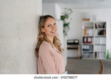 Relaxter junger Büroangestellter, der sich gegen eine Säulenwende zurücklehnt und die Kamera mit einem freundlichen Lächeln in einem geräumigen Firmenbüro mit Kopienraum ansieht