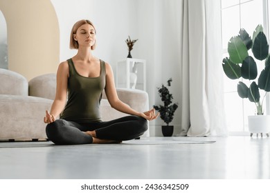 Relaxed young woman doing sitting in yoga position at home. Female athlete sitting in lotus meditating. Serene people concept. Life balance - Powered by Shutterstock