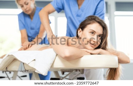 Relaxed young woman lying on couch in spa salon experiencing tandem back massage performed by two expert masseurs ..