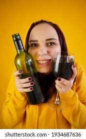 Relaxed Young Woman Poses With Bottle And Glass Of Red Wine On Yellow Background. Adult Happy Brunette Presses Alcohol To Face