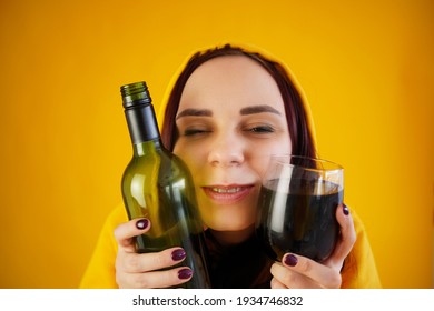 Relaxed Young Woman Poses With Bottle And Glass Of Red Wine On Yellow Background. Adult Happy Brunette Presses Alcohol To Face