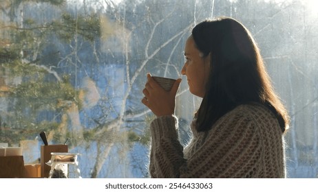 Relaxed young woman sips hot herbal tea in a cafe with views of a snowy forest bathed in winter sunlight - Powered by Shutterstock