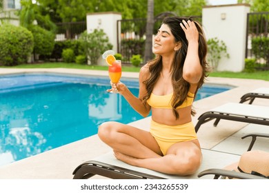 Relaxed young woman smiling while sunbathing by the pool in the summer and enjoying a tropical drink before swimming - Powered by Shutterstock