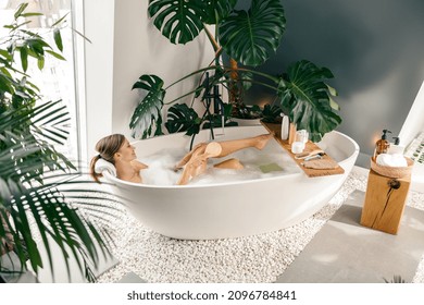 Relaxed Young Woman Using Brush While Taking Buble Bath In Modern Bathroom Decorated With Tropical Plants. Spa, Beauty And Care Concept