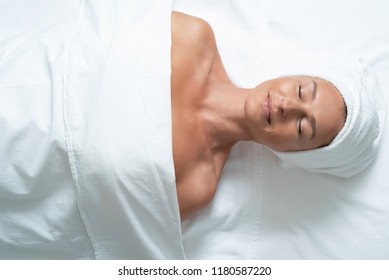 Relaxing After Procedure. Top View Portrait Of Attractive Woman With Closed Eyes Lying Under White Blanket On Massage Table. She Is Wearing Soft Towel On Head