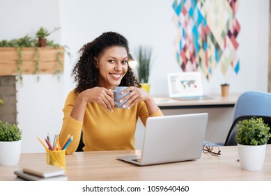 Relaxing. Attractive content young curly-haired woman smiling and drinking tea while working in the office