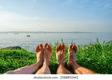 Relaxing by the lakeside, a couple enjoys the peaceful sunny summer view with their bare feet in the grass, overlooking the calm waters and distant horizon - front view