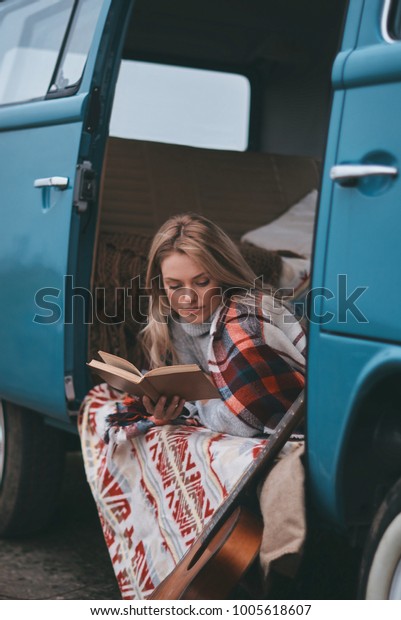Relaxing with her favorite book. Attractive young\
woman covered with blanket reading a book while sitting inside of\
the blue retro style mini\
van