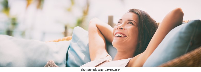 Relaxing home lifestyle happy woman in relax luxury hotel room sofa lying back with arms behind head smiling. Asian girl in comfortable lounging chair travel living.