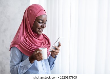 Relaxing Morning. Smiling Black Muslim Woman In Hijab Drinking Coffee And Using Smartphone At Home, Standing Near Window, Enjoying Hot Drink And Mobile Communication, Reading Messages, Copy Space - Powered by Shutterstock