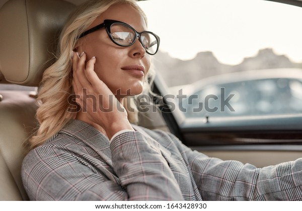 Relaxing on the way to the office.\
Attractive and stylish businesswoman in eyeglasses adjusting\
headphones while driving a car. Transportation.\
Music