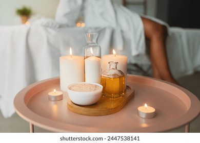 Relaxing spa setting with lit candles, massage oil, and a bowl on a pink side table over black lady in white bathrobe - Powered by Shutterstock