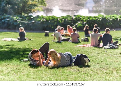 Relaxing Summer Day In A Danish Park, People Lounging On The Grass, Pond Water In The Background. Concept Of Hygge.