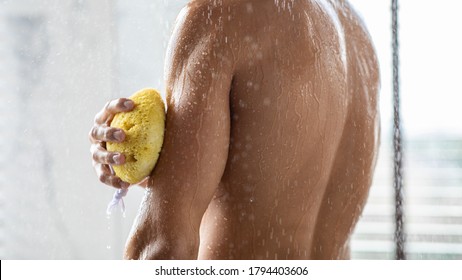 Relaxing Time Concept. Muscular man taking shower and washing his arms and shoulders with sponge, back view
