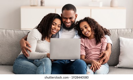 Relaxing Together. Portrait of happy African American parents and their cute daughter using laptop, spending time together, watching movie or surfing internet, sitting on the sofa in living room