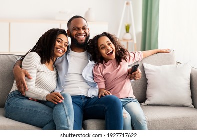 Relaxing Together. Portrait of happy young African American family watching television, parents sitting with their daughter on comfortable sofa in the living room, laughing at the program