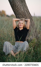 Relaxing Under A Tree Blond Woman Sitting On Grass. She Is Wearing Skirt Pants And Black Shirt. Her Eyes Closed, Hands On Back Of Her Head. Low Angle.