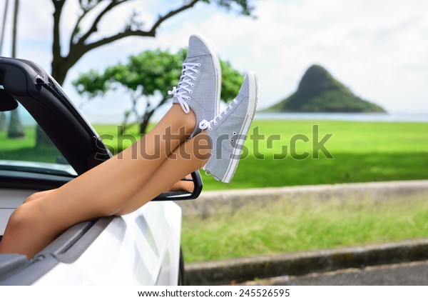 Relaxing vacation car road\
trip travel with feet up the window. Convertible car holiday\
concept with female legs in front of Oahu landmark : Chinaman\'s Hat\
in Hawaii, USA.
