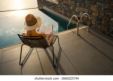 relaxing vacation - woman sitting in chair and reading a book on terrace near swimming pool at luxury villa