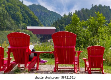 Relaxing Woman, Reading A Book,sitting On Red Muskoka  Lounging Chair, With A Wonderful View, Of The Point Wolfe Covered Bridge  In Fundy National Park, New Brunswick Canada