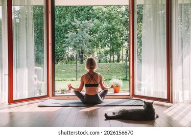 Relaxing Woman Sitting In A Lotus Position In Front Of The Opened Window, While Doing Yoga At Home. Her Cat Is Watching Her, Laying Down Next To Her.