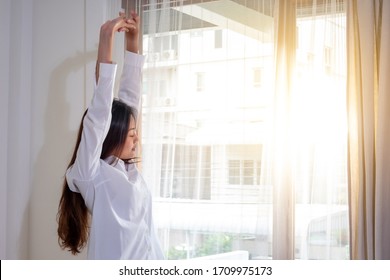 Relaxing Young Beautiful Asian Woman Girl Stretching After Wake Up In Bedroom Near The Window With Sun Shining Through The Curtain Into The Room In The Morning. Healthy Sleeping And Wellbeing Concept