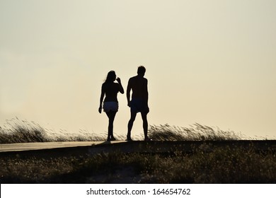 Relaxing Young Couple Silhouette. Man And Woman Walking By A Pathway On A Beach.