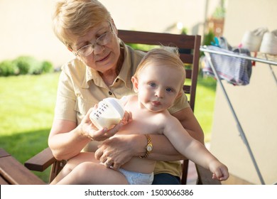 Reliable Baby Sitter. Granny Taking Care Baby. Feeding Baby With Bottle. Grandmother Enjoying The Feeding Of Her New Baby Boy Grand Sun Feeling Proud. Help To Young Mother