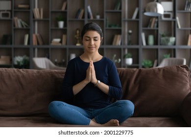 Relieving stress. Relaxed millennial indian woman sit on sofa alone in lotus position clasp hands in namaste. Serene young lady meditate at home breath deep with closed eyes feel harmony peace of mind