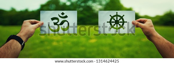 Religion conflicts as global issue concept. Two\
hands holding different faith symbols, Buddhism vs Hinduism belief\
over green field nature. Relations between different people\
doctrines and cult.