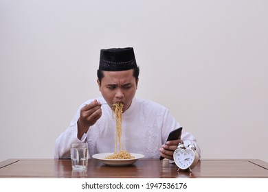 Religious Asian Man In Koko Shirt Or White Muslim Shirt And Black Cap Eating Instant Noodles For Suhoor To Be Strong During Fasting From Dawn Until Dusk In Ramadan Month While Using Mobile Phone